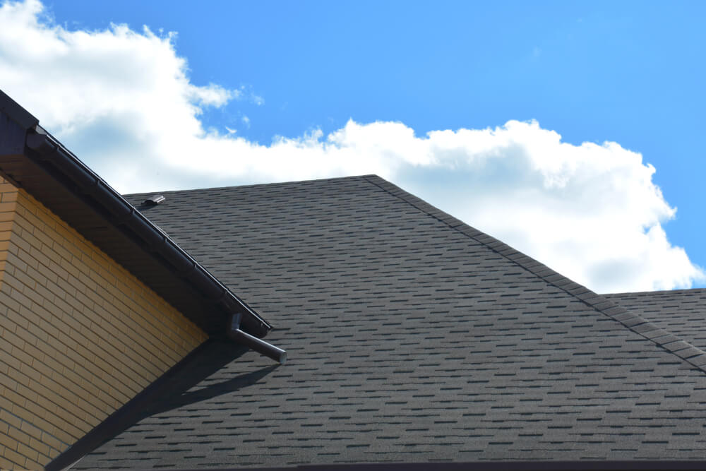 A pitched roof of a residential building with gray asphalt shingles and visible guttering, under a blue sky with scattered white clouds, prompts the curious to ask: what is a hip roof?