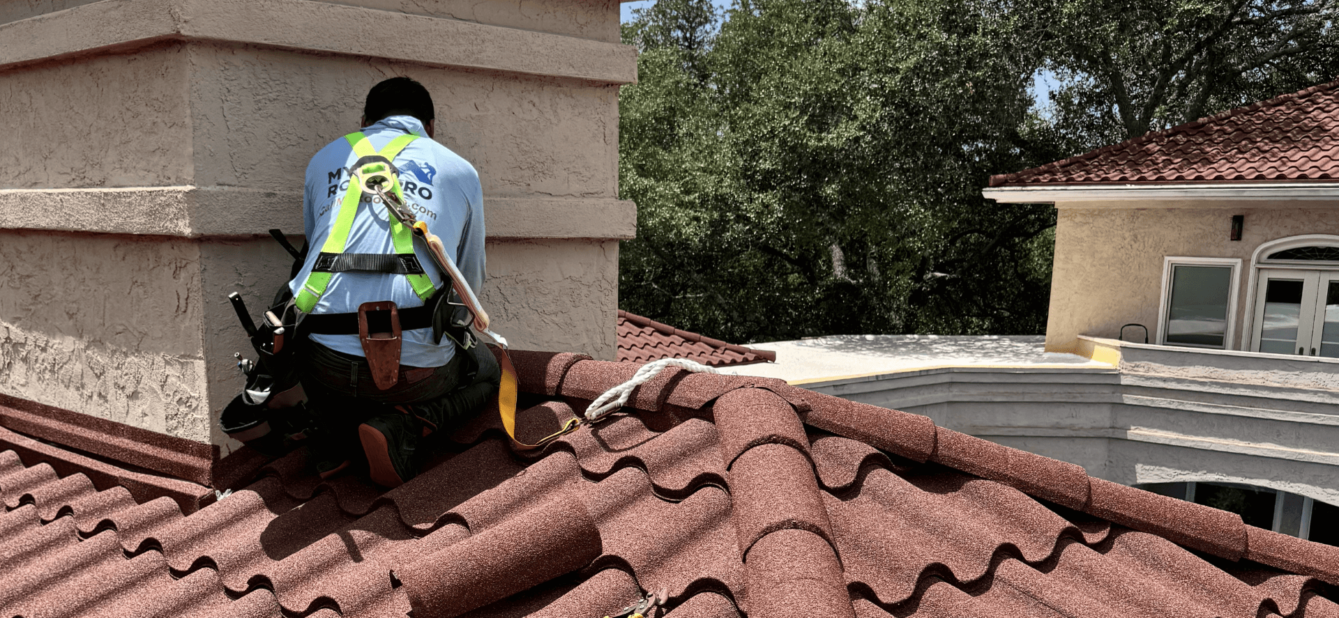 Roofing professional inspects chimney on red tile roof with safety harness.