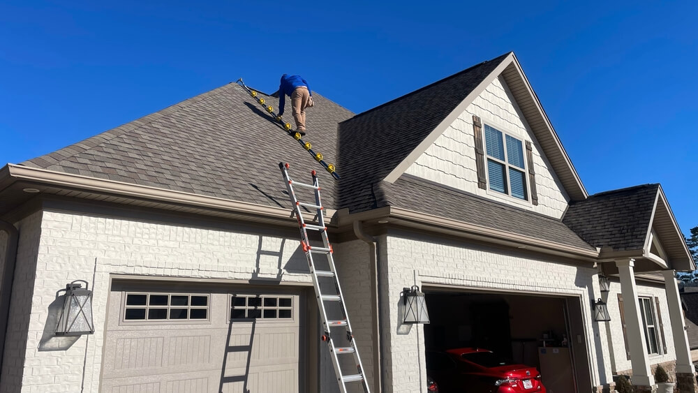 Roof technician adjusting ladder; another ladder on garage. Clear sky backdrop.